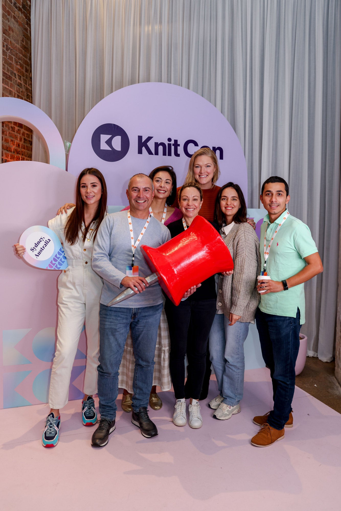 A group of people pose in an auditorium with a giant papier-mâché pin prop. 